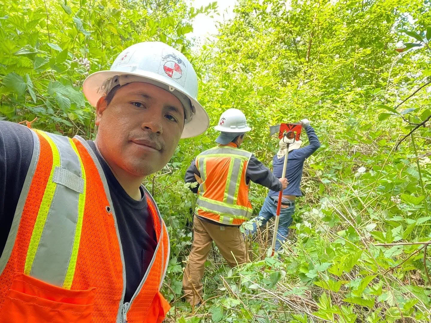 Survey crew in hard hats and high-visibility vests working through heavy brush with a prism pole during a topographic survey.
