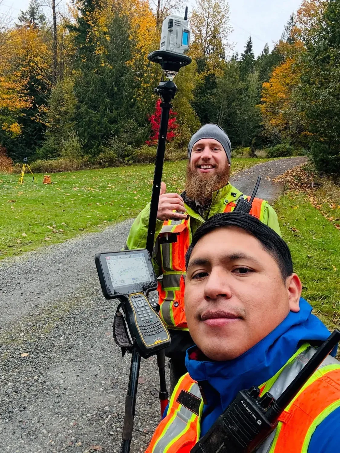 Two surveyors in high-visibility gear with a GNSS RTK rover and data collector on a rural access road.